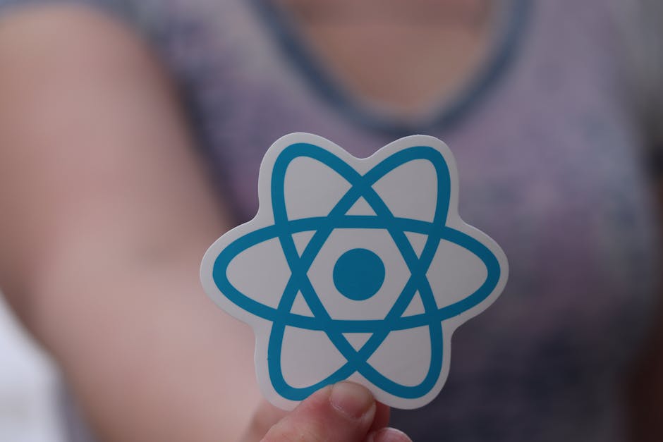 Close-up of a person holding a white sticker with a blue atomic symbol, featuring an orbiting electrons diagram, against a blurred background of a person wearing a purple and gray top inside a room. T