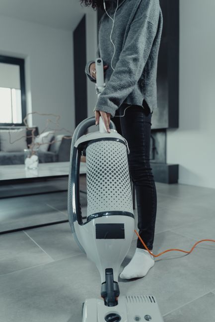 Close-up of a person vacuuming a wooden floor with a modern canister vacuum cleaner, which features a black wand and hose connected to a transparent dust container. The individual is wearing grey shor