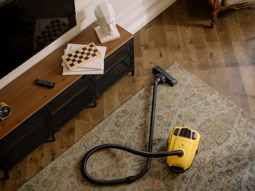 A woman performing surface cleaning with a yellow and black vacuum cleaner on a detailed patterned area rug in a living room. The rug features floral motifs in shades of beige, light green, and orange