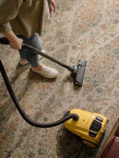 A person kneeling on a patterned Victorian-style carpet in a living room, preparing a yellow and black portable vacuum cleaner for surface cleaning. The individual is wearing a tan jacket and blue jea